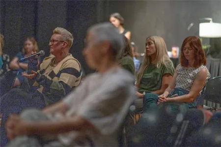 Audience members listen and engage in discussion during an indoor event or seminar, with a person speaking into a microphone.