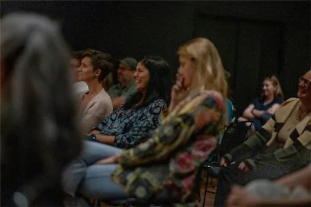 Audience members seated and smiling during an engaging event or presentation, captured in a dimly lit room.