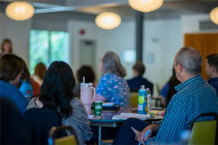 People attending a seminar, sitting around tables with notebooks and water bottles in a brightly lit room.