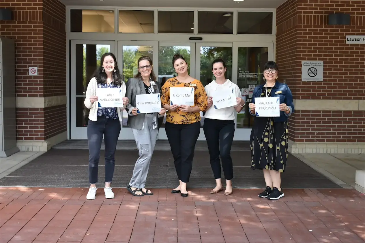 Five people holding multilingual welcome signs outside a building.