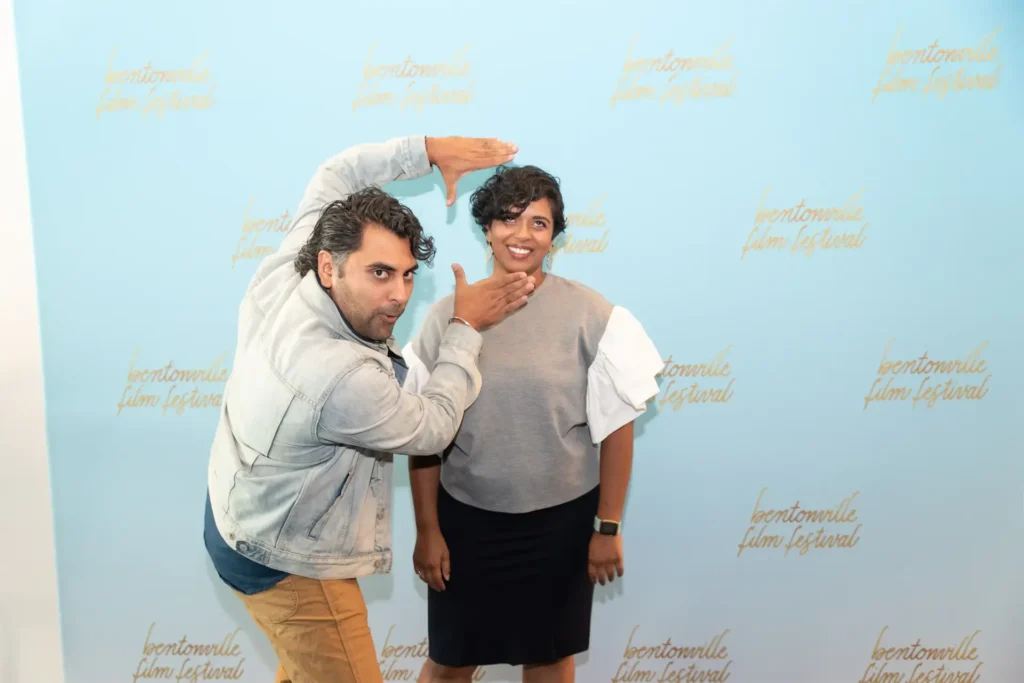 Two people pose playfully at the Bentonville Film Festival, with a light blue backdrop and festival logo.