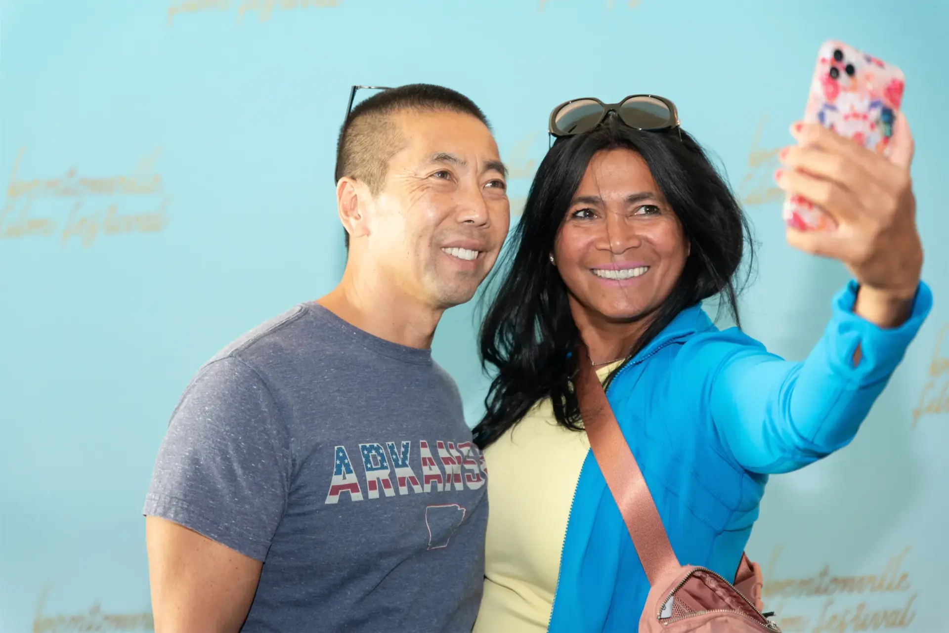 Smiling couple taking a selfie against a light blue backdrop at an event.