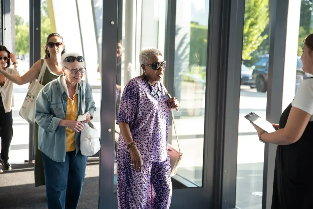 Group of diverse people walking through a glass entrance on a sunny day, wearing casual clothing and sunglasses.