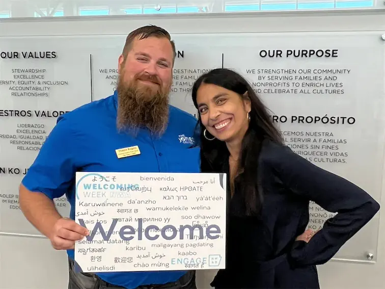 Two people holding a multilingual Welcome sign, standing in front of a board displaying organization values.
