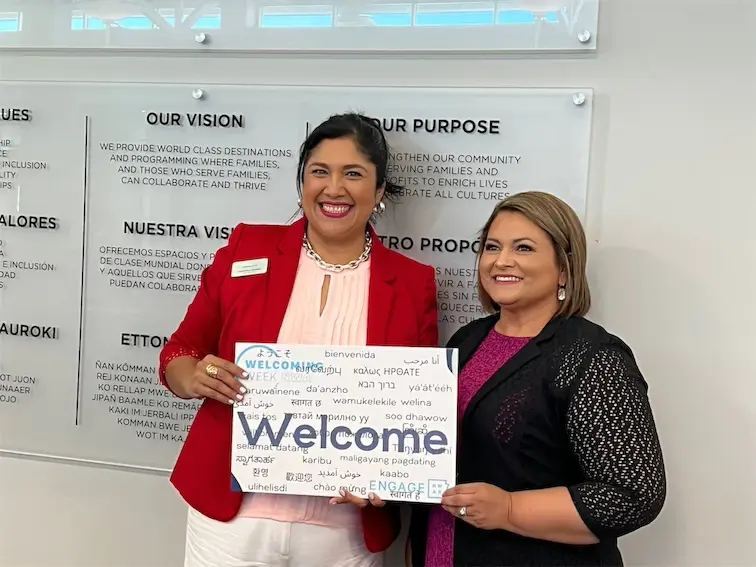 Two individuals smiling, holding a multilingual Welcome sign in front of values and purpose statement on a wall.
