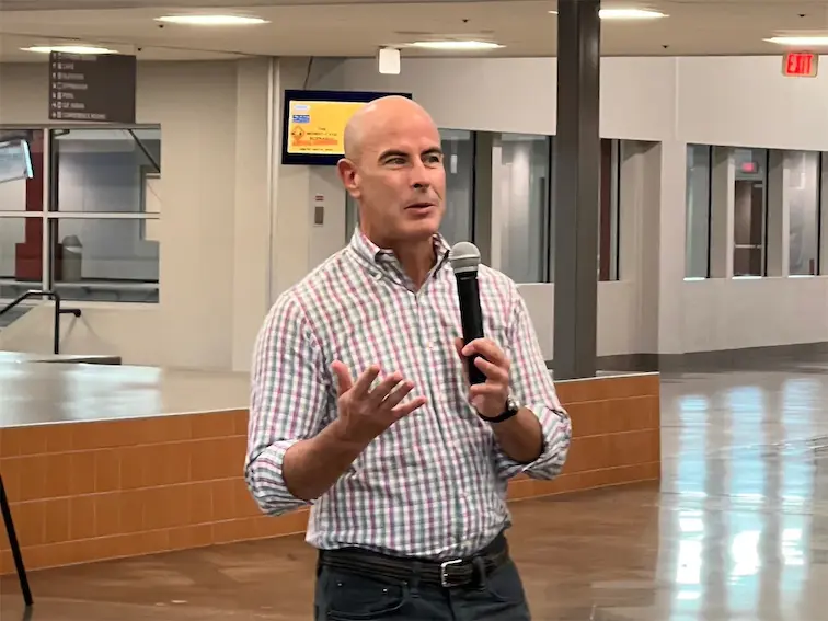 Man speaking into a microphone in an indoor setting, wearing a checkered shirt.