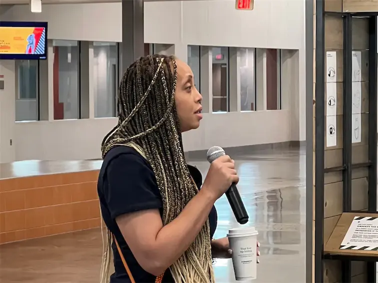 Person speaking into a microphone indoors, holding a coffee cup, with a modern office background.