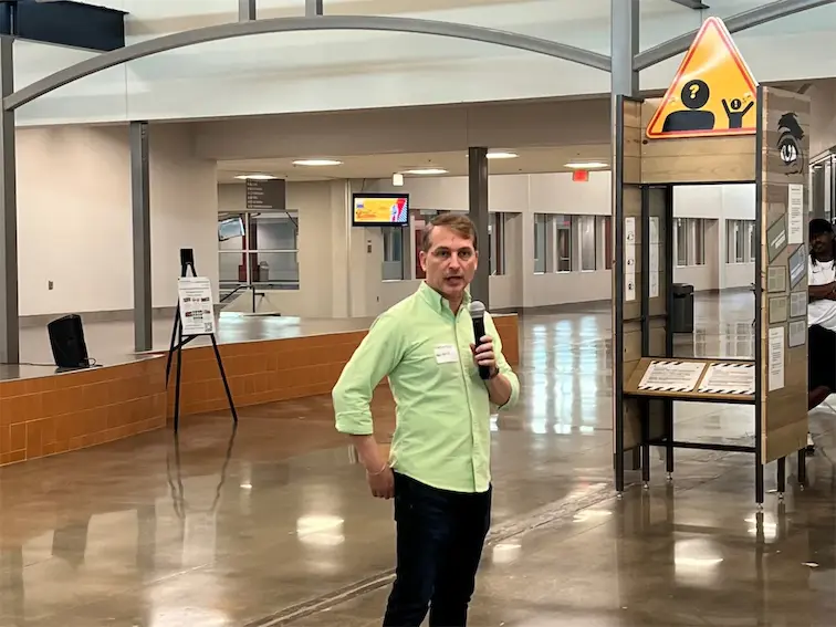 Person speaking with a microphone in a modern, open venue with informational signs in the background.