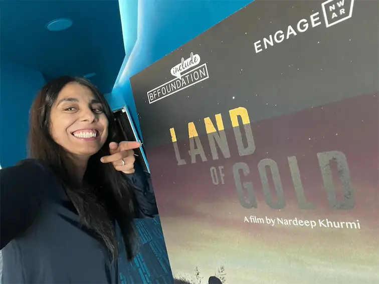 Smiling person pointing at Land of Gold film poster by Nardeep Khurmi in a bright blue setting.
