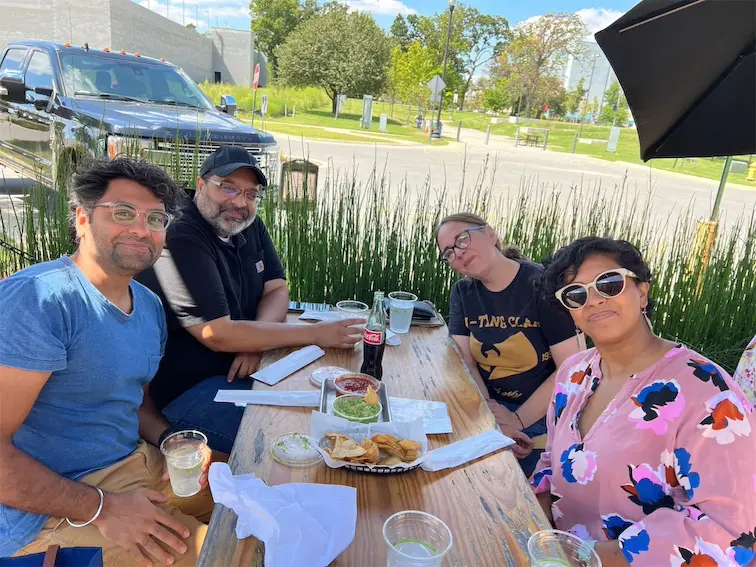 Group enjoying a sunny outdoor meal with drinks and snacks at a wooden table, green plants in the background.