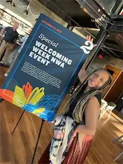 Person smiling next to a Welcoming Week NWA Event sign in an indoor venue.
