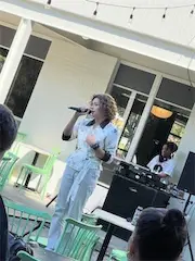 Singer performing on outdoor stage with a microphone, DJ in background, green chairs in audience area.