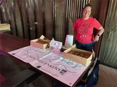 Person standing at a bakery stall with cookies displayed, featuring a pink banner and a casual, inviting setup.