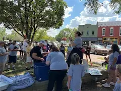 Community event with people gathering in a park under trees, colorful buildings in the background on a sunny day.