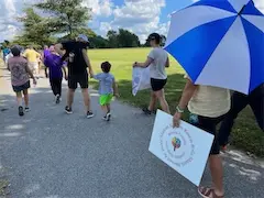 Community group walking on a sunny path, some carrying signs and umbrellas, with a grassy field in the background.