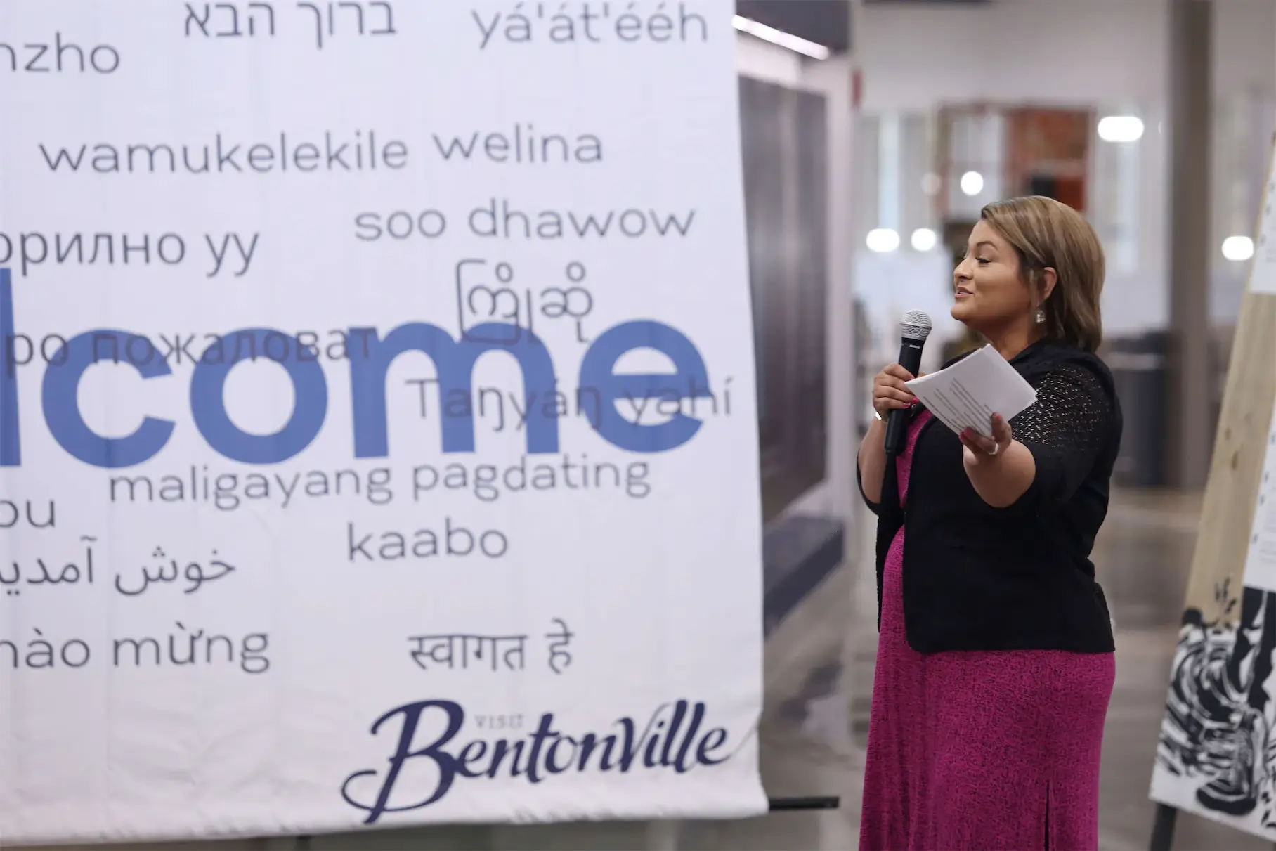 Woman speaking at a multilingual welcome event in Bentonville with a large banner in the background.