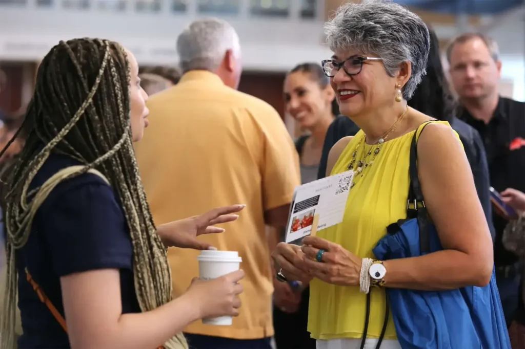 Two women engaging in conversation at a social event, one holding a coffee cup and the other a brochure.