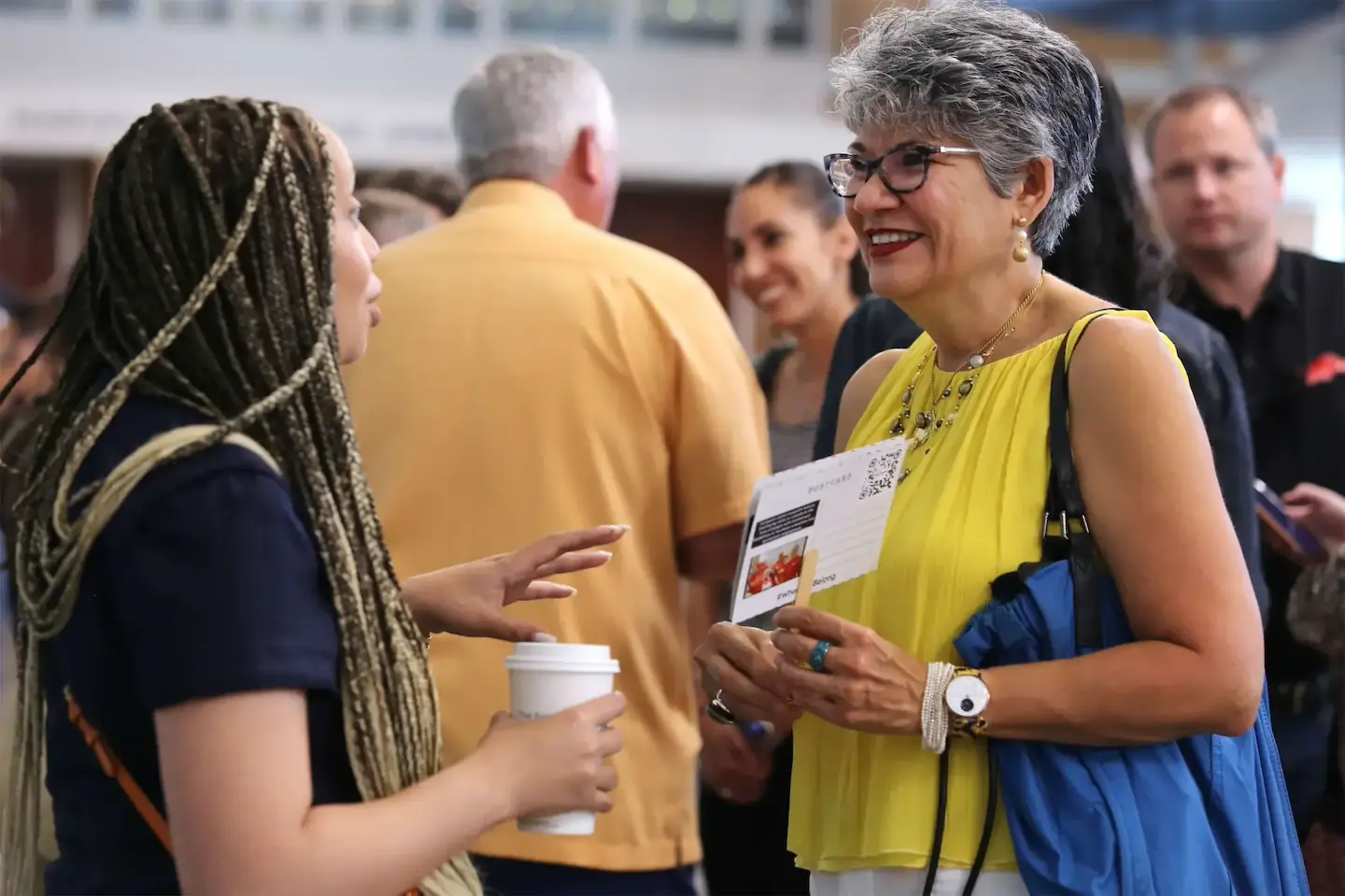 Two women engaging in conversation at a social event, one holding a coffee cup and the other a brochure.