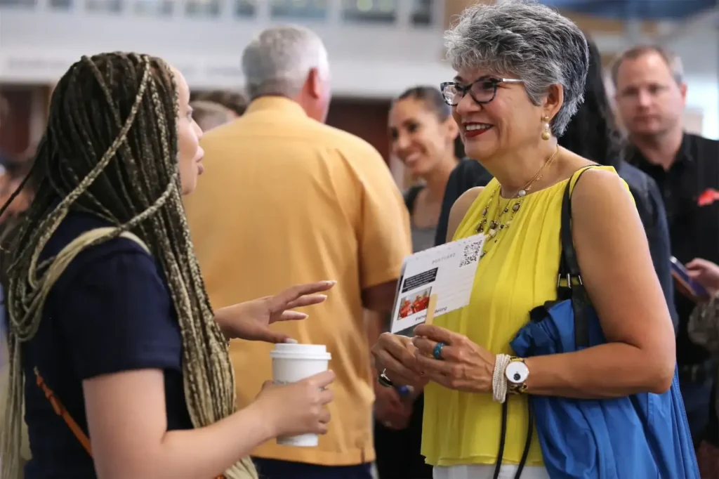 Two women engaging in lively conversation at a networking event, one holding a coffee, the other smiling with papers.