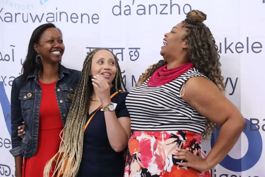 Three women smiling and laughing in front of a banner with text, wearing colorful and casual outfits.