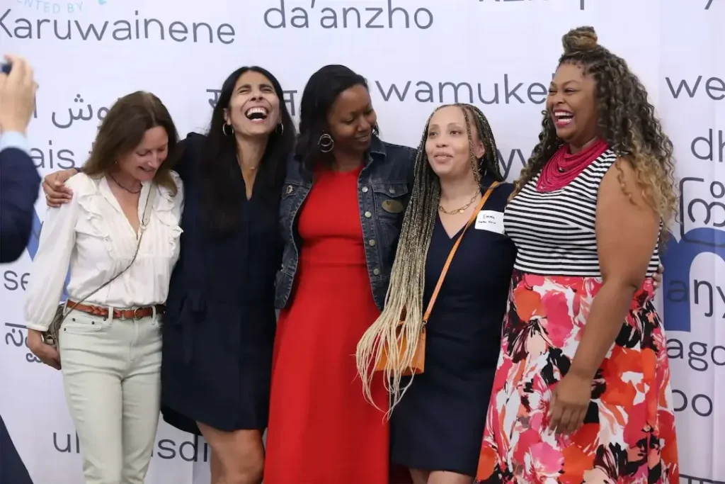 A group of diverse women laughing together in front of a multilingual background at an event.