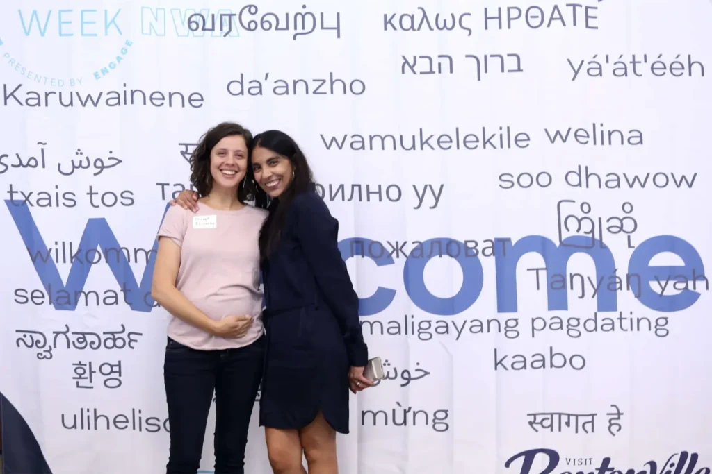 Two smiling women standing together against a multilingual Welcome backdrop at an event.
