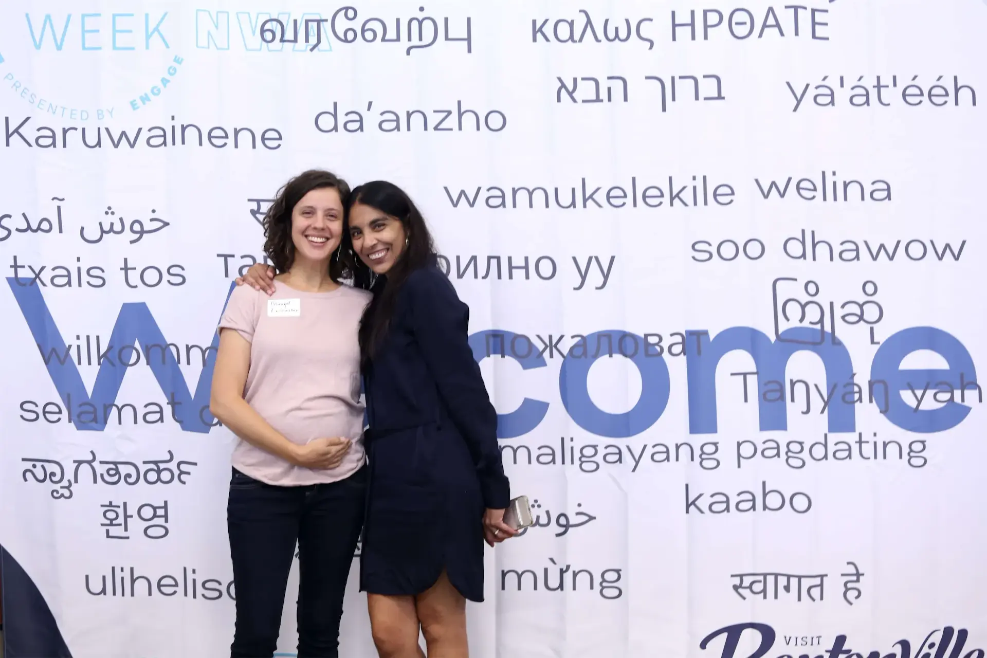Two smiling women standing together against a multilingual Welcome backdrop at an event.