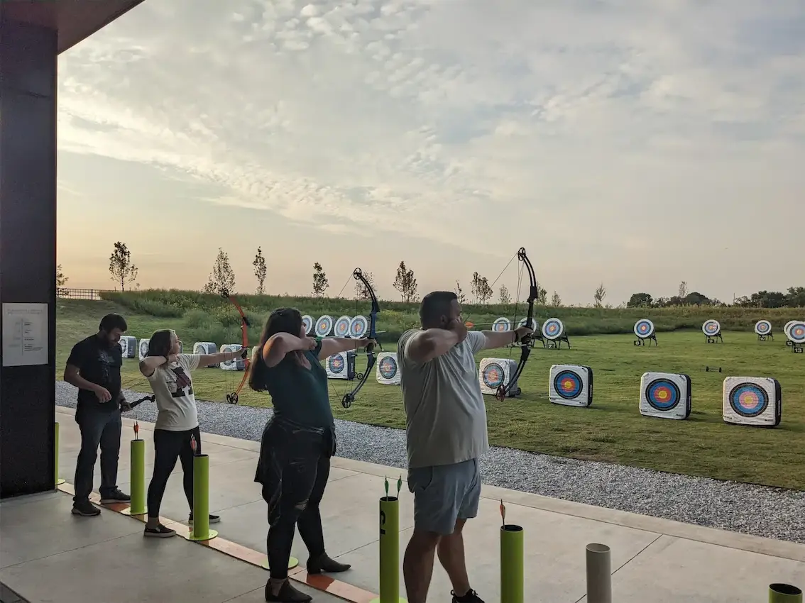 People practicing archery at a range, aiming at targets against a scenic outdoor backdrop.