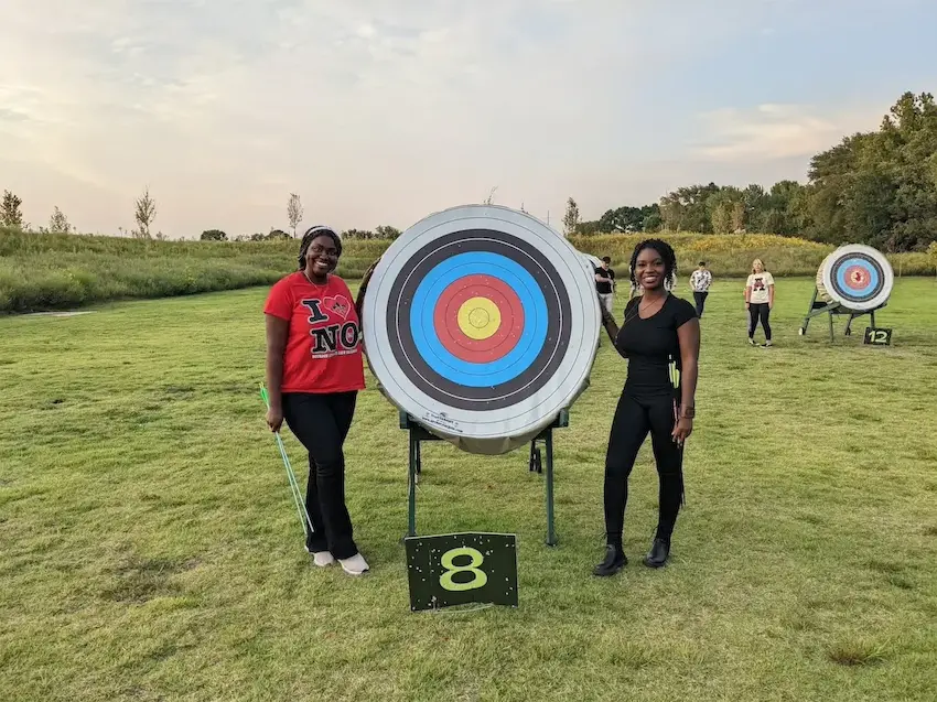 Two women smiling next to an archery target on a grassy field, with more targets and people in the background.