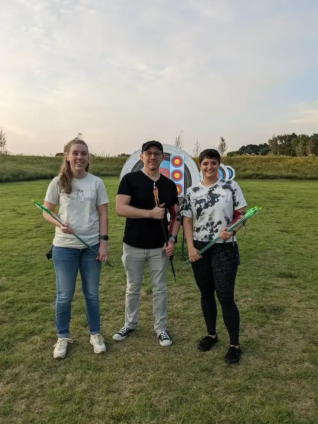 Three people standing in a field with archery targets and arrows, smiling outdoors.