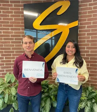 Two people holding Bienvenida signs in front of a brick wall with a stylized 'S' logo, welcoming visitors.