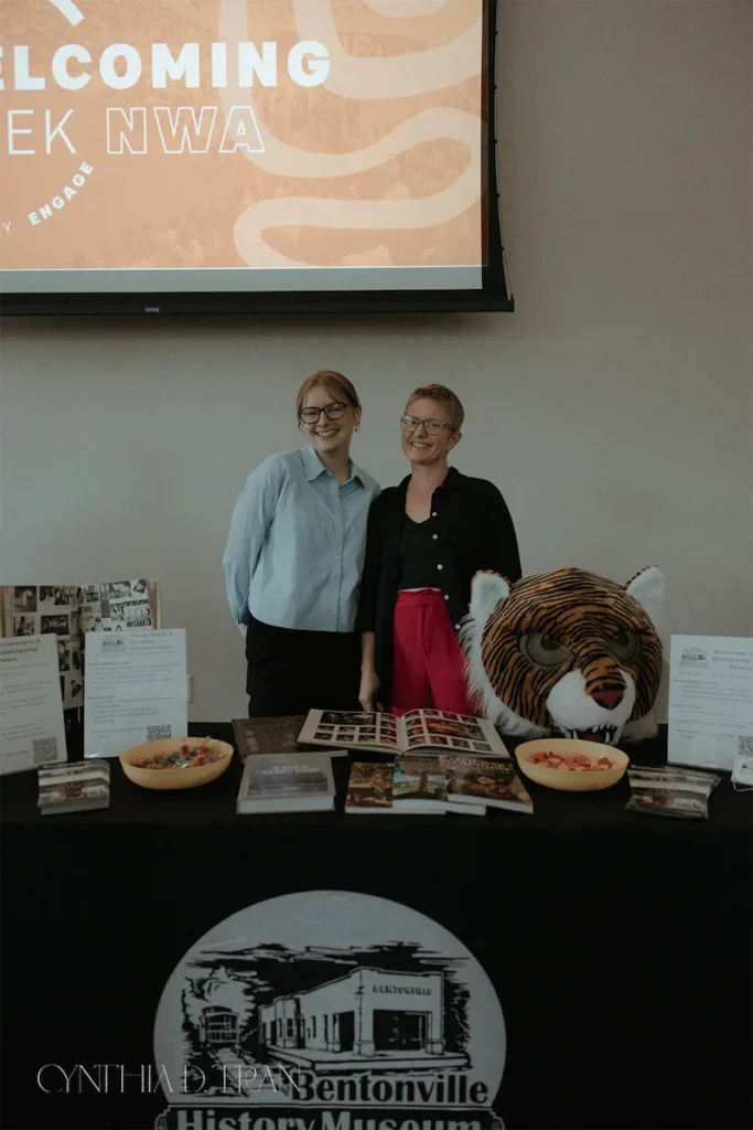 Two people smiling at a museum event table with brochures and a tiger mascot head.