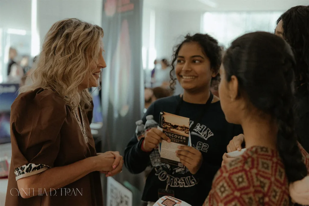 A woman talks to smiling attendees at an indoor event, exchanging flyers and enjoying the conversation.