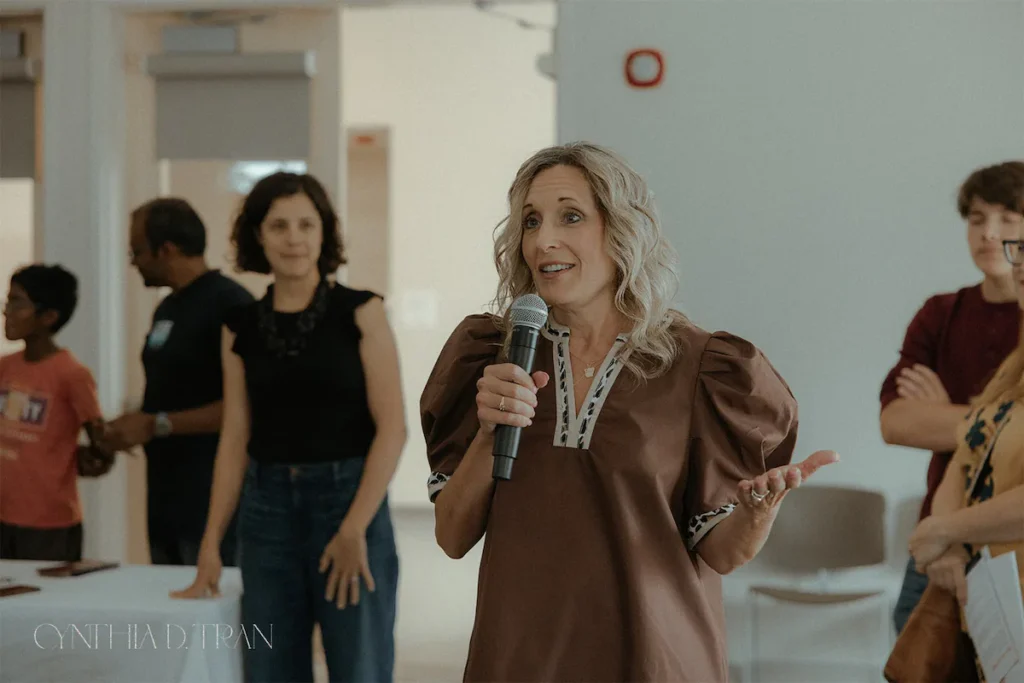 Speaker addressing an audience at an indoor event, holding a microphone and gesturing with one hand.