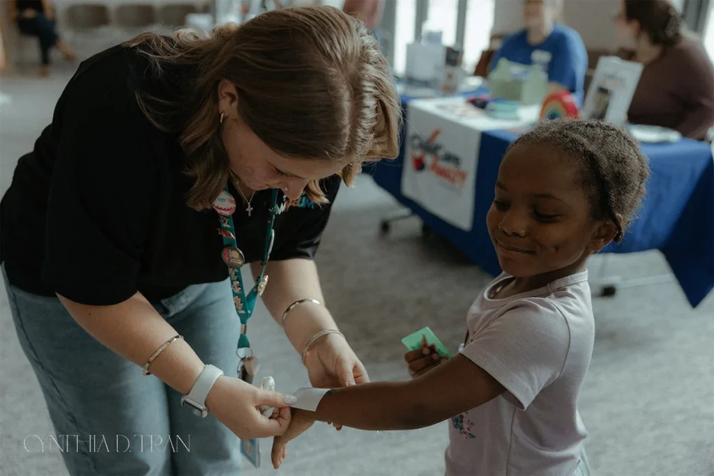 Woman helping child with wristband at an event, smiling, indoor setting with informational table in background.