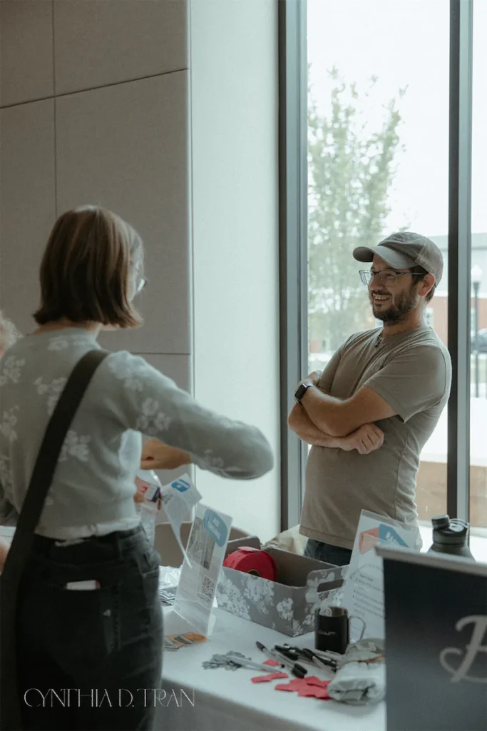 Two people conversing at an event table near a window, surrounded by promotional materials and supplies.