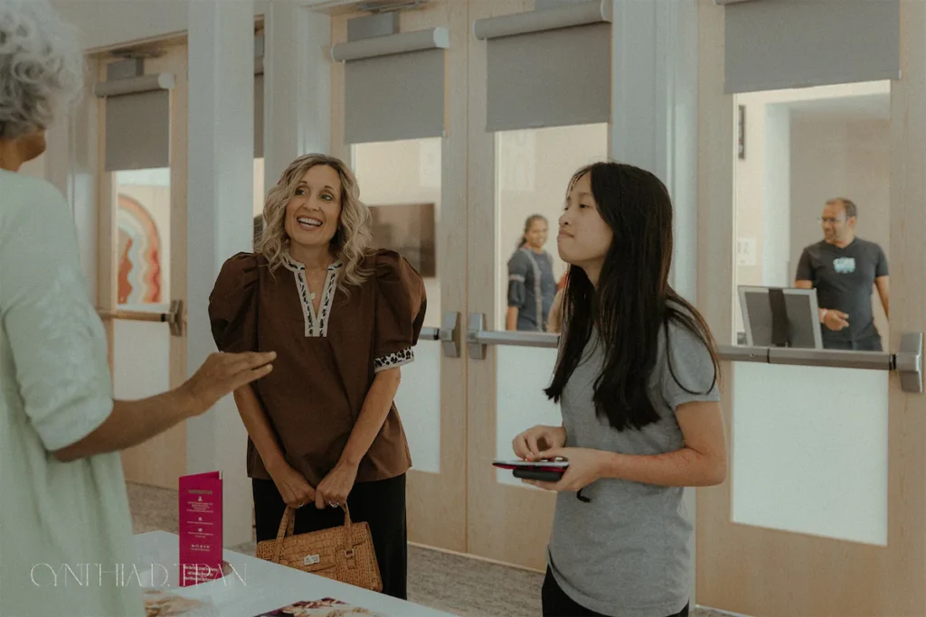 Women interacting warmly at a community event, with one holding a phone near a table.