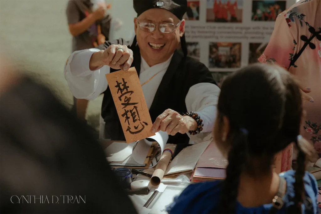 Smiling person holds up calligraphy art at a cultural event, engaging with a crowd.