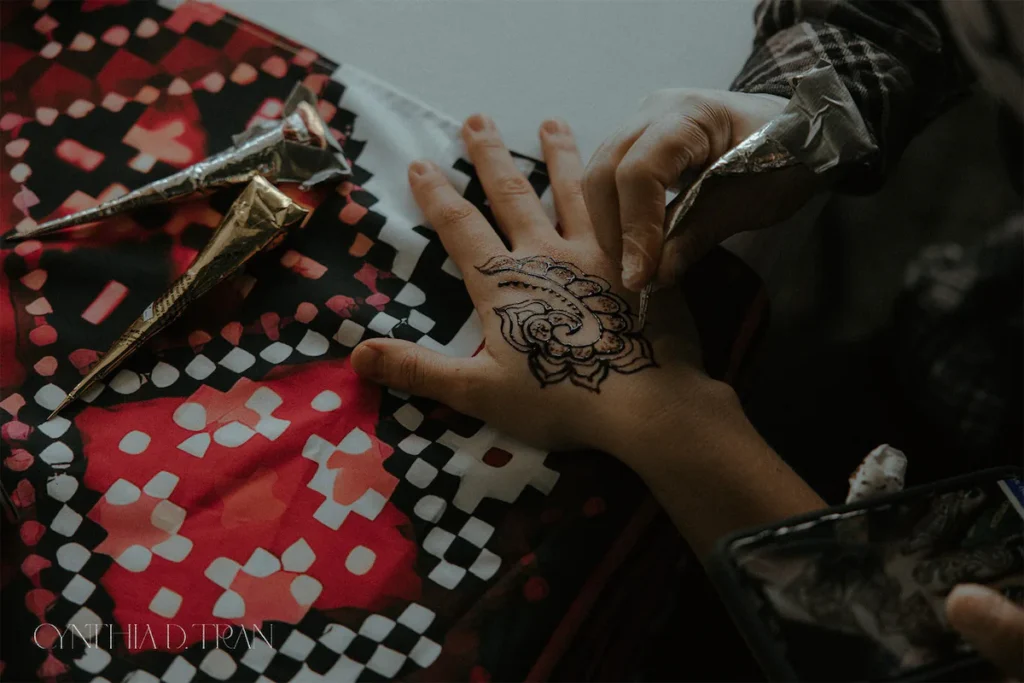 Hand being decorated with intricate henna design, surrounded by colorful patterned fabric.