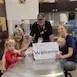 Group of people smiling in a kitchen holding a Welcome sign, symbolizing hospitality and community gathering.